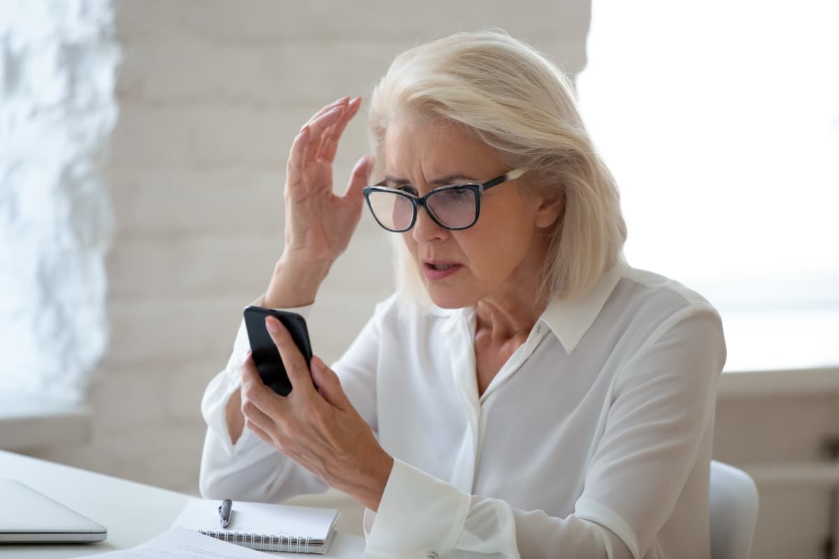 Confused senior businesswoman sit at office desk hold cellphone