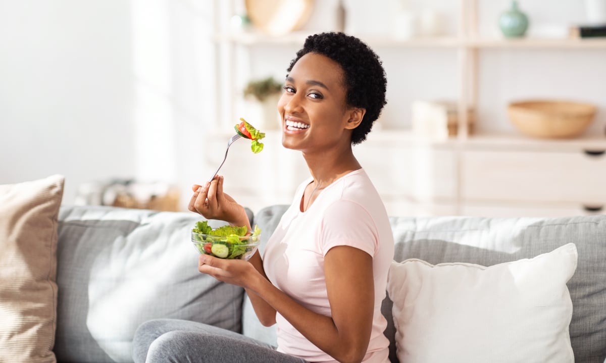 Healthy nutrition concept. Pretty black woman eating yummy vegetable salad on sofa