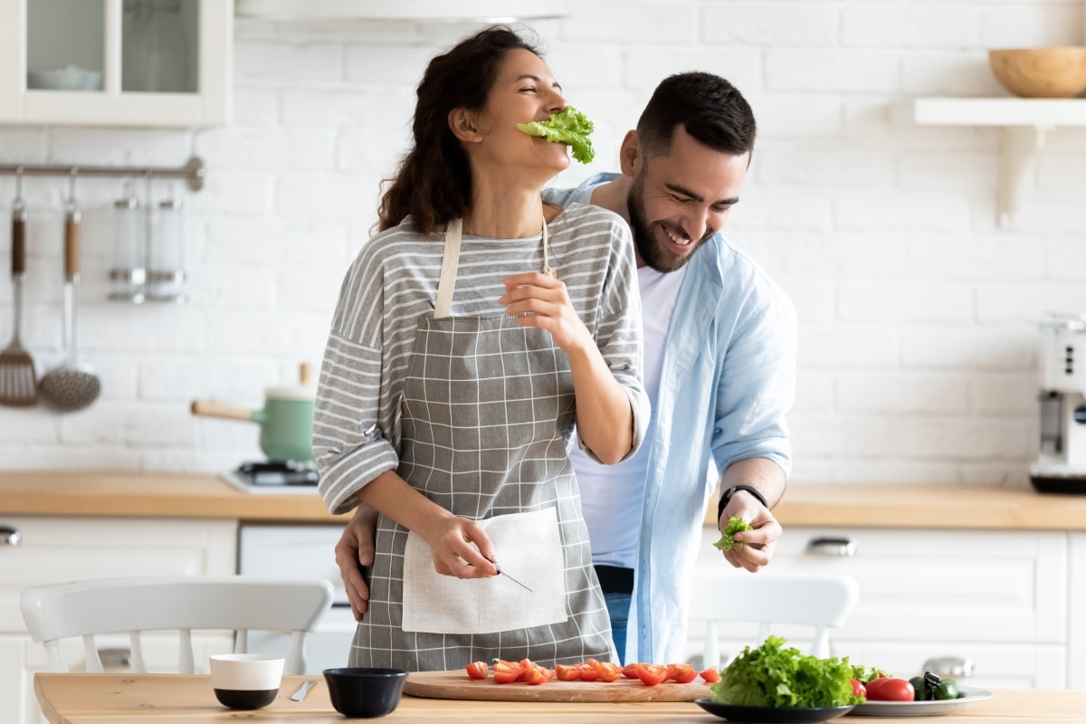 Millennial couple preparing healthy vegetarian salad laughing enjoying process and time with better half on modern cozy kitchen, cooking together during first dating