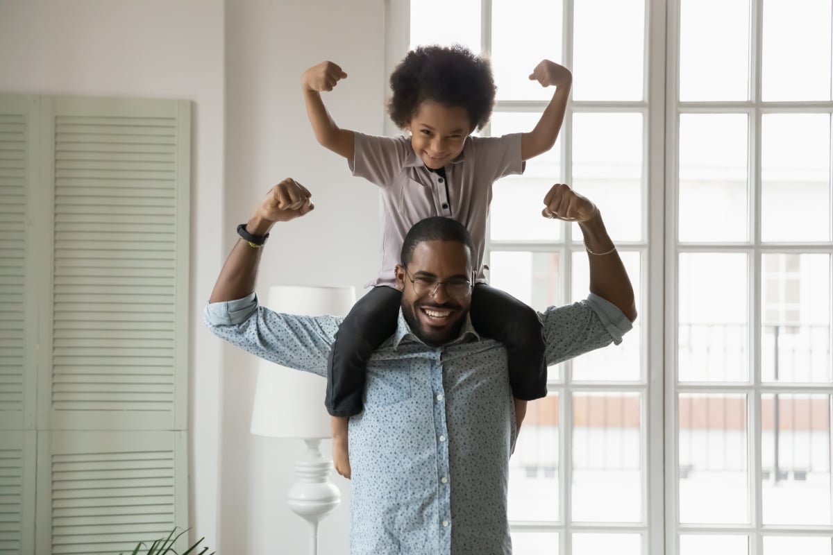 Small son sit on strong dad shoulders showing biceps. African family enjoy activity games at home