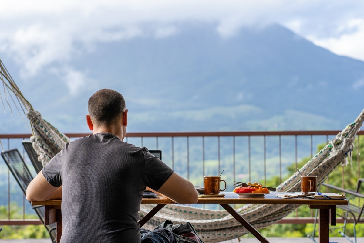 Man sitting on a table with breakfast while working with a view of a volcano in front of him. Costa Rica