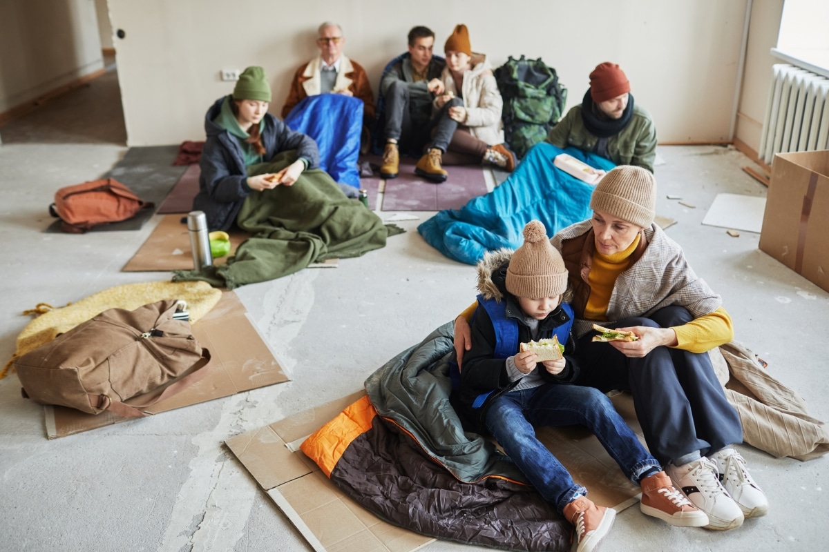 High angle view at group of Caucasian refugees eating food while hiding in shelter on floor covered with blankets