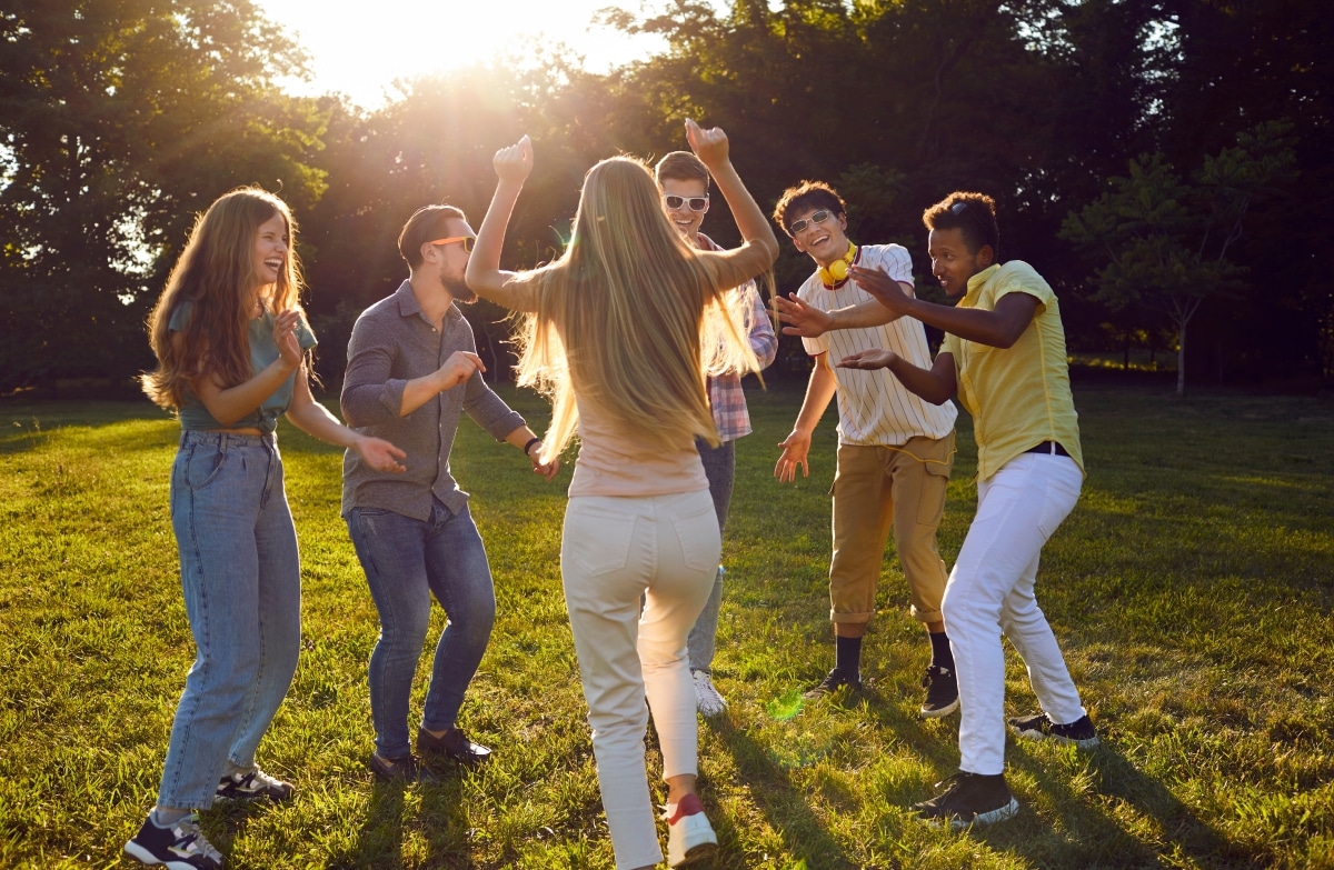 Funny group of young friends having fun dancing on green lawn in park on summer evening. Friendship concept.
