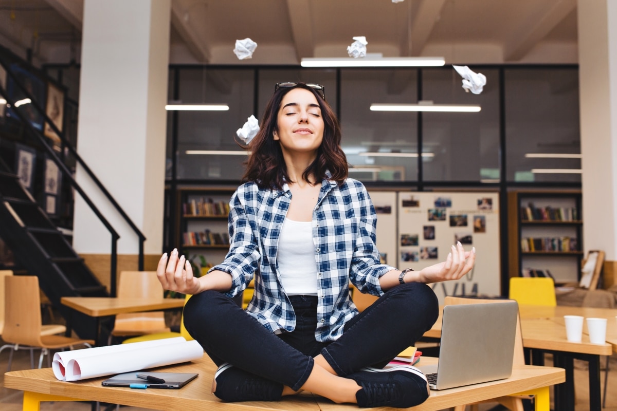 Young joyful brunette woman having meditation on table surround work stuff and flying papers. Cheerful mood
