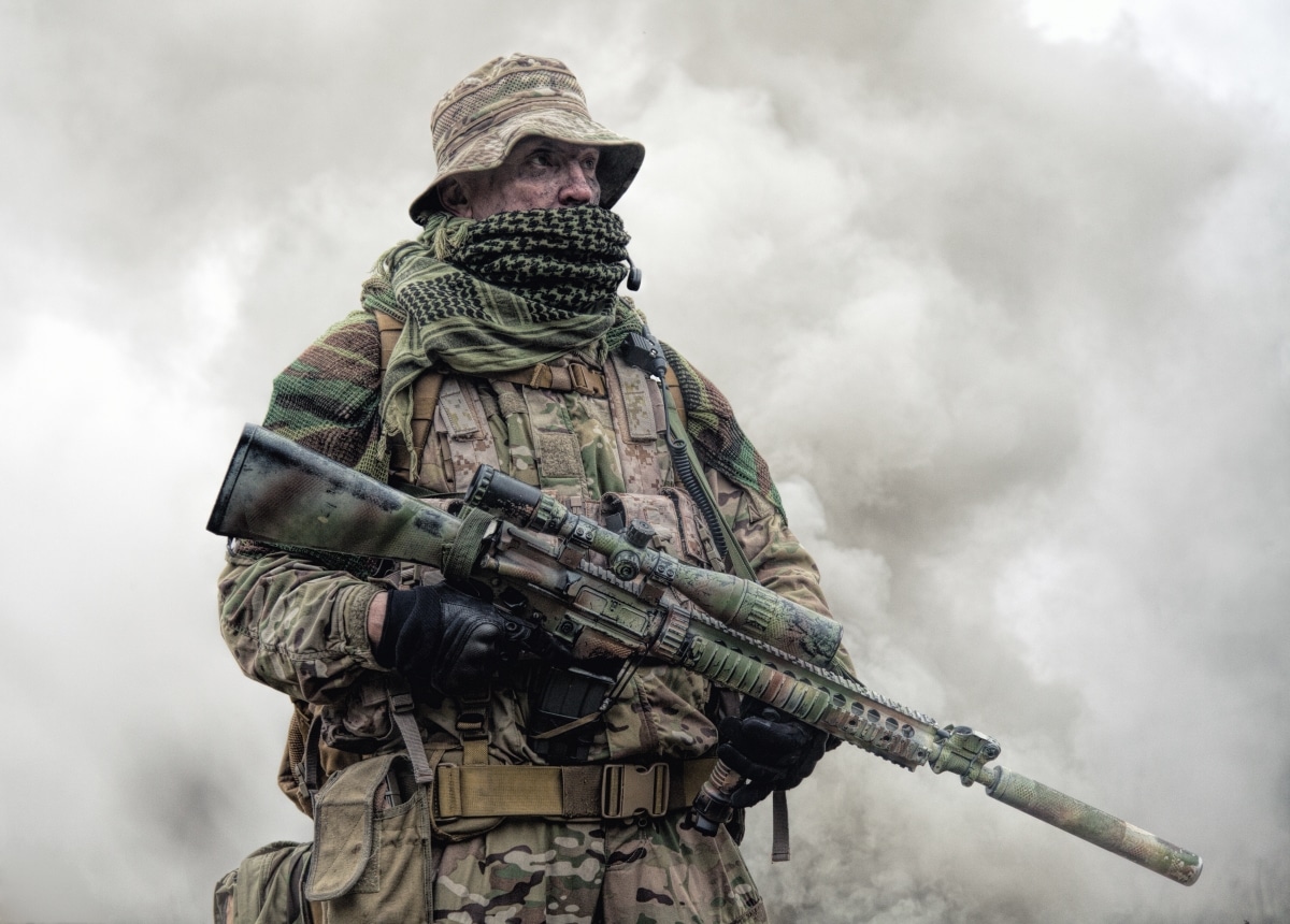 Elite commando fighter, private military company mercenary, army special forces veteran in camouflage uniform, shemagh and bonnie, equipped radio headset standing with sniper rifle in clouds of smoke