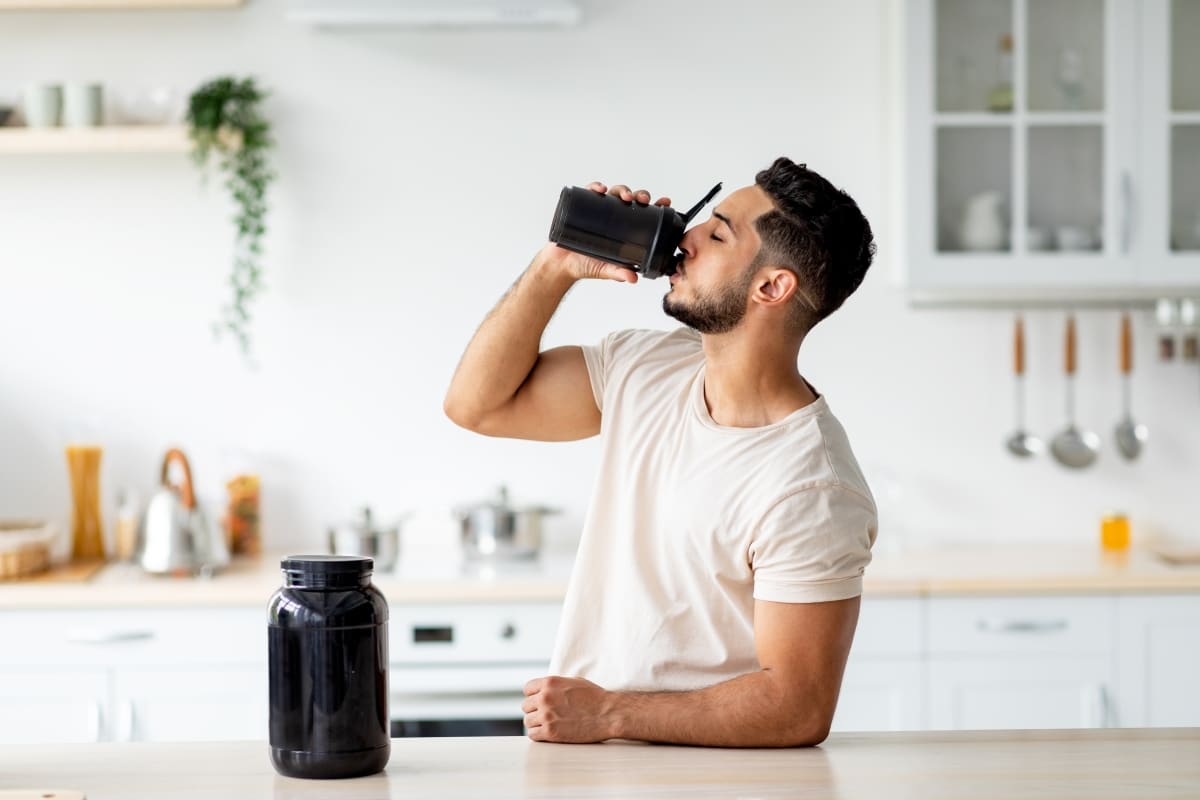 Young Arab guy drinking protein shake from bottle at kitchen, copy space.