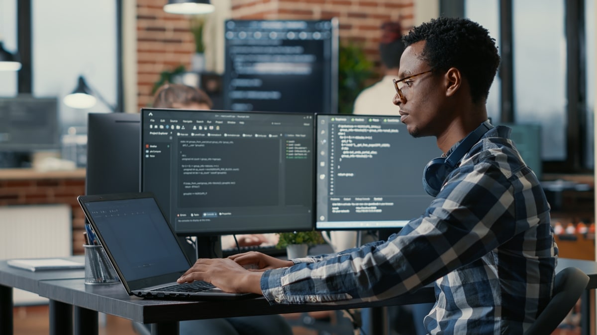 Portrait of african american developer using laptop to write code sitting at desk with multiple screens parsing algorithm in software agency. Coder working on user interface using portable computer.