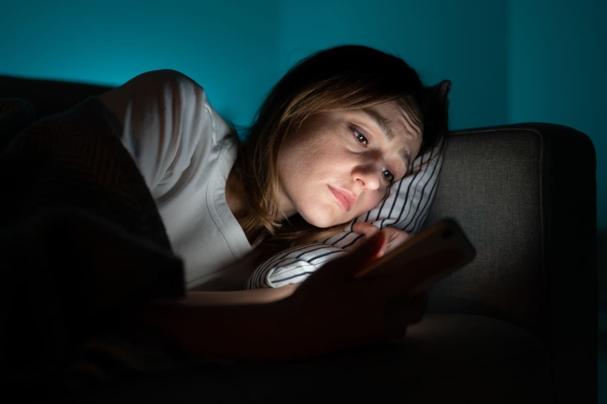 Unhappy woman lying in bed with smartphone, following ex-boyfriend on social media, sad female using mobile phone at night, can not sleep after relationship breakup