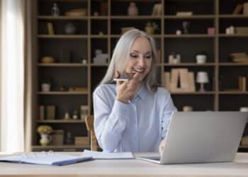 Middle-aged businesslady holds smartphone talks on speakerphone sit at desk, leave voice message via messenger, text on laptop.