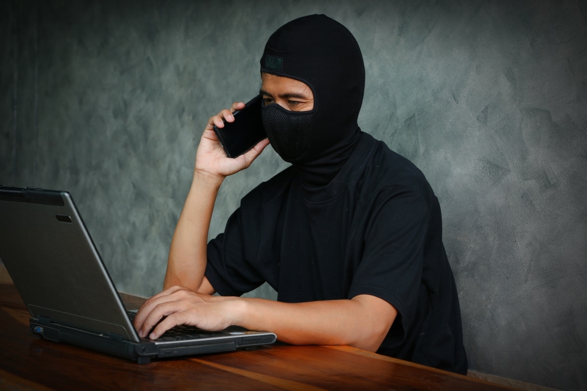 Male hacker in a robber mask uses phone, and laptop in some fraudulent scheme.