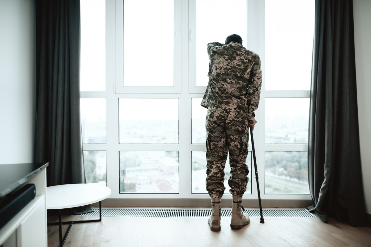 A disabled soldier stands with his hand on the window. He feels sad and depressed. It stands near a large panoramic window, from which you can see the city.