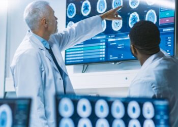Team of Scientists Work in the Brain Research Laboratory, Discussing Brain Scans Show on Wall TV Monitor. Neurologists / Neuroscientists Surrounded by Monitors Showing CT, MRI Scans.
