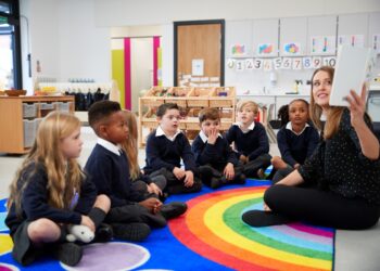 Female teacher holding up a book in front of her class of elementary school kids sitting on the floor in a classroom, side view