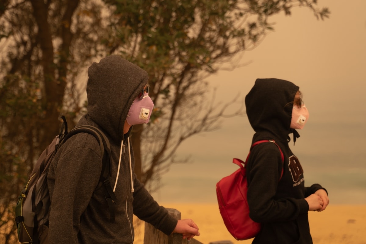 Eden, Australia - December 31, 2019: Tourists, a father and daughter at the beach in Eden by the evacuation center, wearing particulate masks and sun glasses to avoid the dense orange bushfire haze.