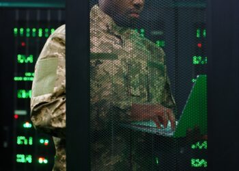 Close up of young African American military officer checking work of servers in analytic monioring center. Man with laptop computer control networking in dark room Cybersecurity in army concept.