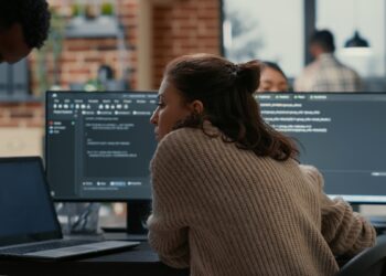 Software programer putting laptop with source code on colleague coder desk asking for opinion about database. Developer writing algorithm interrupted by coworker wanting help with fixing errors.