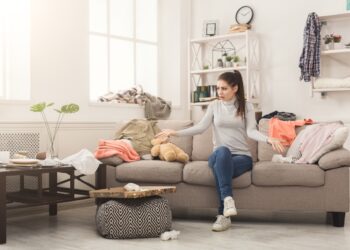 Desperate helpless woman sitting on sofa in messy living room. Young girl surrounded by many stack of clothes. Disorder and mess at home, copy space