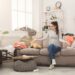 Desperate helpless woman sitting on sofa in messy living room. Young girl surrounded by many stack of clothes. Disorder and mess at home, copy space