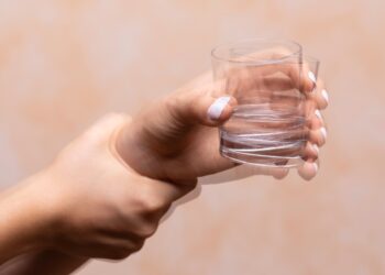 A closeup view on the hands of a person trying to hold a glass of water steady, shaking hands symptomatic of a central nervous and motor system disease such as Parkinson's