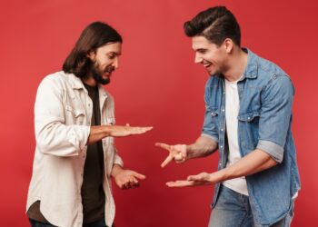 Photo of young smiling guys playing rock-paper-scissors game and laughing isolated over red background
