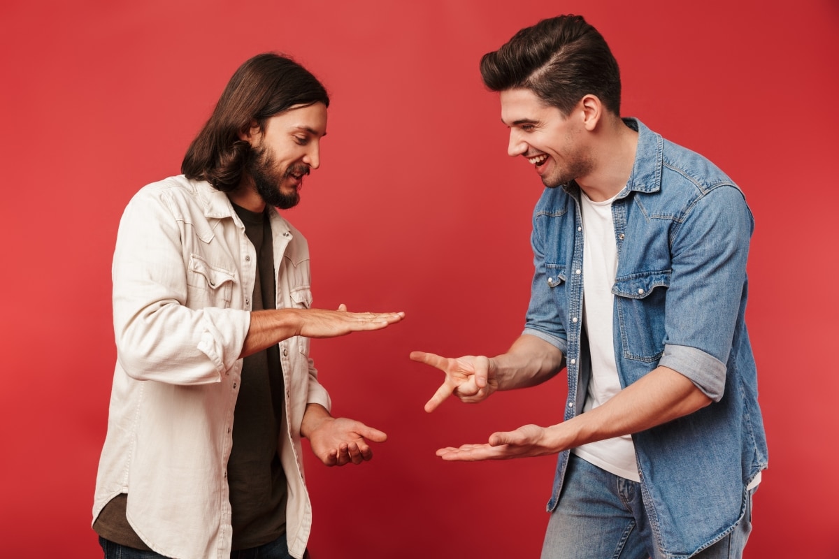 Photo of young smiling guys playing rock-paper-scissors game and laughing isolated over red background