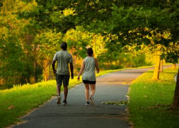Image of an African American man and a caucasian woman walking on a hiking trail. The couple wears matching vests, shirts, shorts and sneakers. These two people walk peacefully on a sunny day