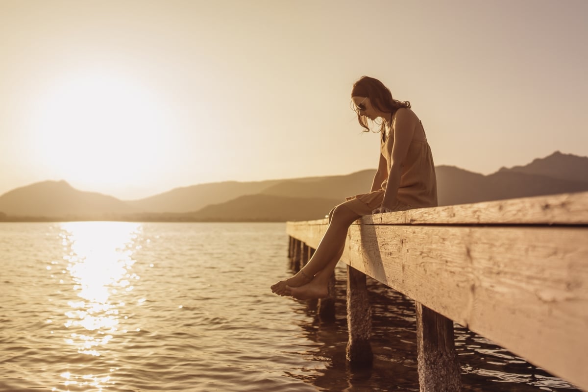 Pensive single Caucasian pretty young woman sitting on a pier of a lake looking down to the water at sunset - Vintage orange color mood