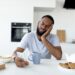 Apathy. Portrait of tired sleepy black man using smartphone while having breakfast in kitchen at home, bored African American male sitting at table, reading text messages on his cell phone eating food