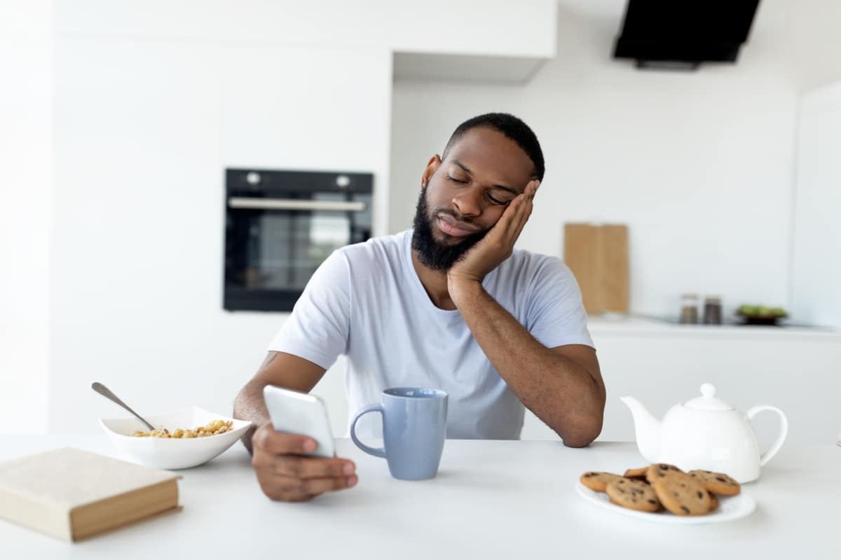 Apathy. Portrait of tired sleepy black man using smartphone while having breakfast in kitchen at home, bored African American male sitting at table, reading text messages on his cell phone eating food