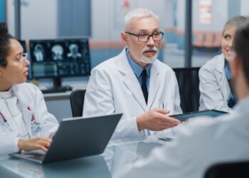 Hospital Conference Meeting Room: Senior Neuroscientist Uses Tablet Computer with MRI Magnetic Resonance Image of the Brain, Talks to Team of Medical Doctors, Neurologists Discuss Patient Treatment