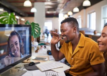 A Black man waves to his colleague on a video call from his office