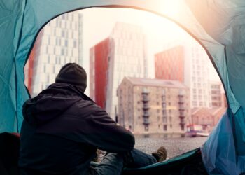 View from inside of a homeless tent. Man in dark dirty cloths sitting by the entrance looking at rich high value district houses. Living during financial crisis concept. Dreaming on better future.