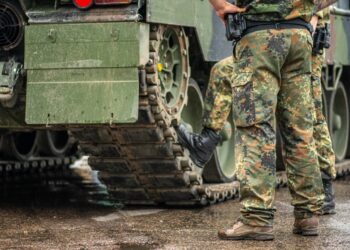 Two soldiers in camouflage uniform standing during a break under the rain near the tracks of an armoured tank, only the trousers and the boots are visible