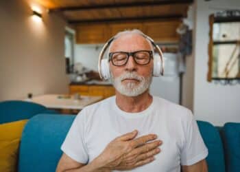 One man senior caucasian male eyes closed for guided training yoga or meditation while sitting at home with headphones self-care practice real people well-being inner peace and balance concept