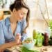 Young and happy woman eating healthy salad sitting on the table with green fresh ingredients indoors