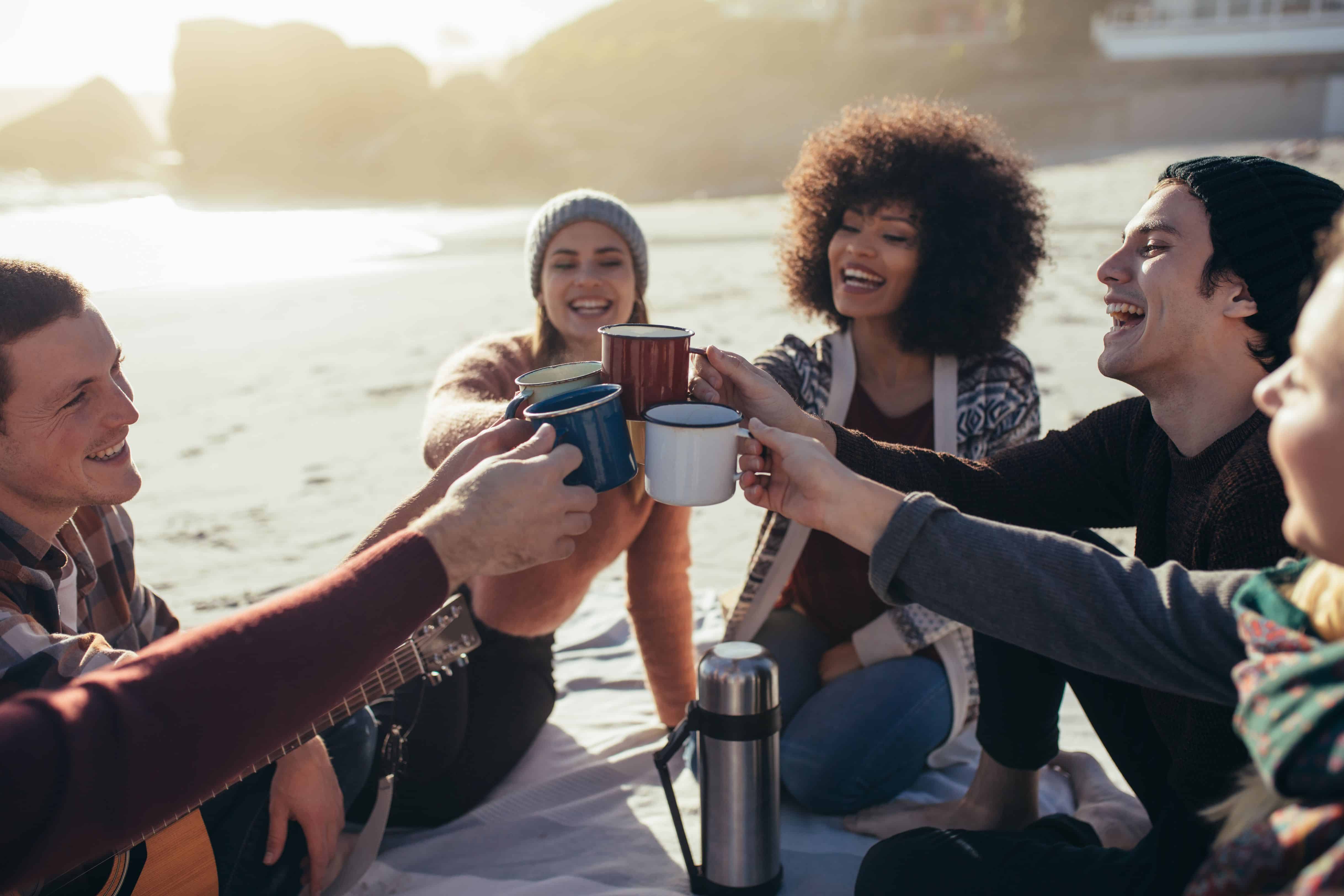 Group of multi-ethnic young people toasting coffee mugs on the beach. Group of friends spending time together at the beach having coffee.