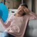 Overheated female sitting on couch in living room at hot summer weather day feeling discomfort suffers from heat waving blue fan to cool herself, girl sweating dwelling without air conditioner concept