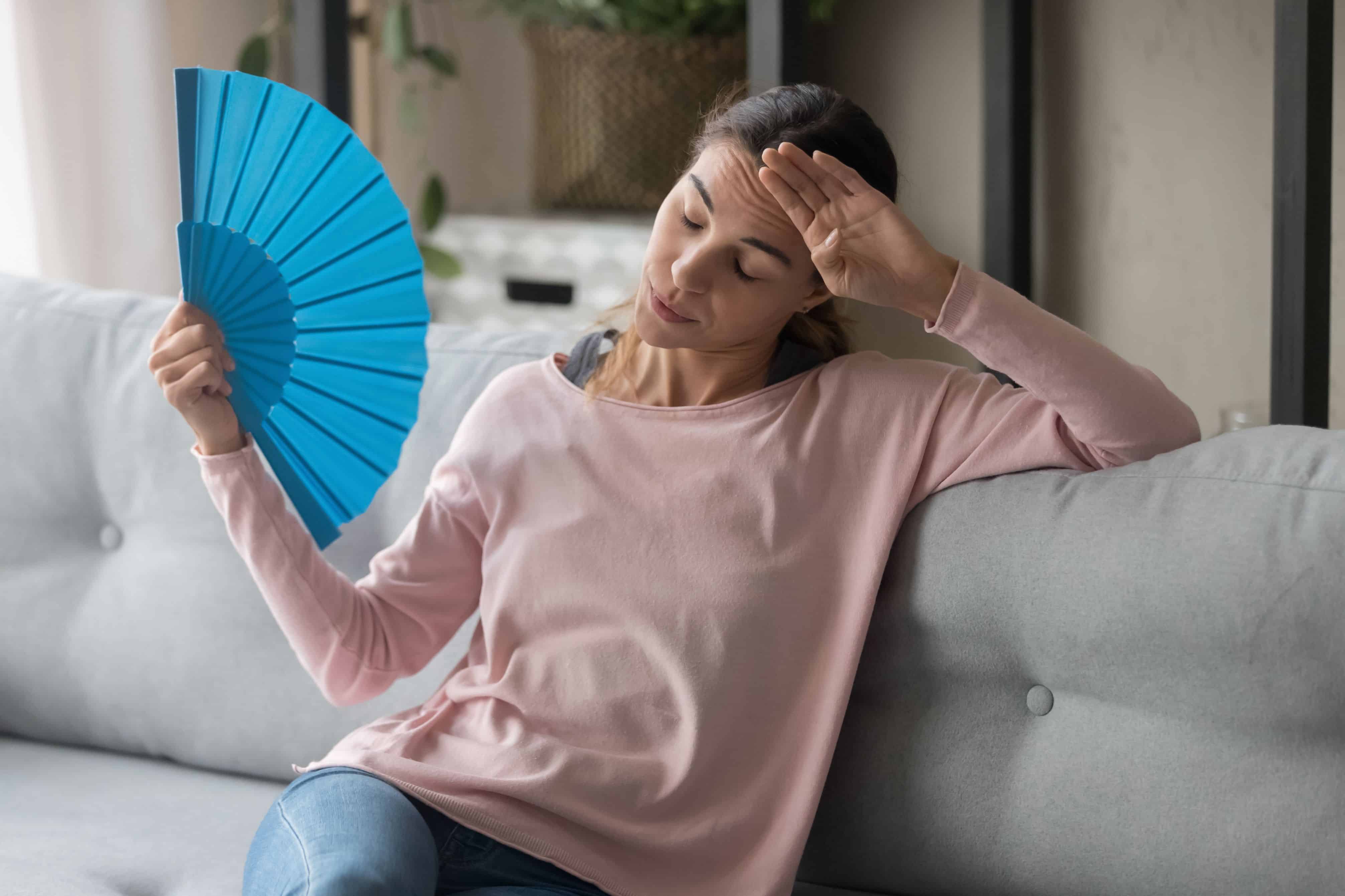 Overheated female sitting on couch in living room at hot summer weather day feeling discomfort suffers from heat waving blue fan to cool herself, girl sweating dwelling without air conditioner concept