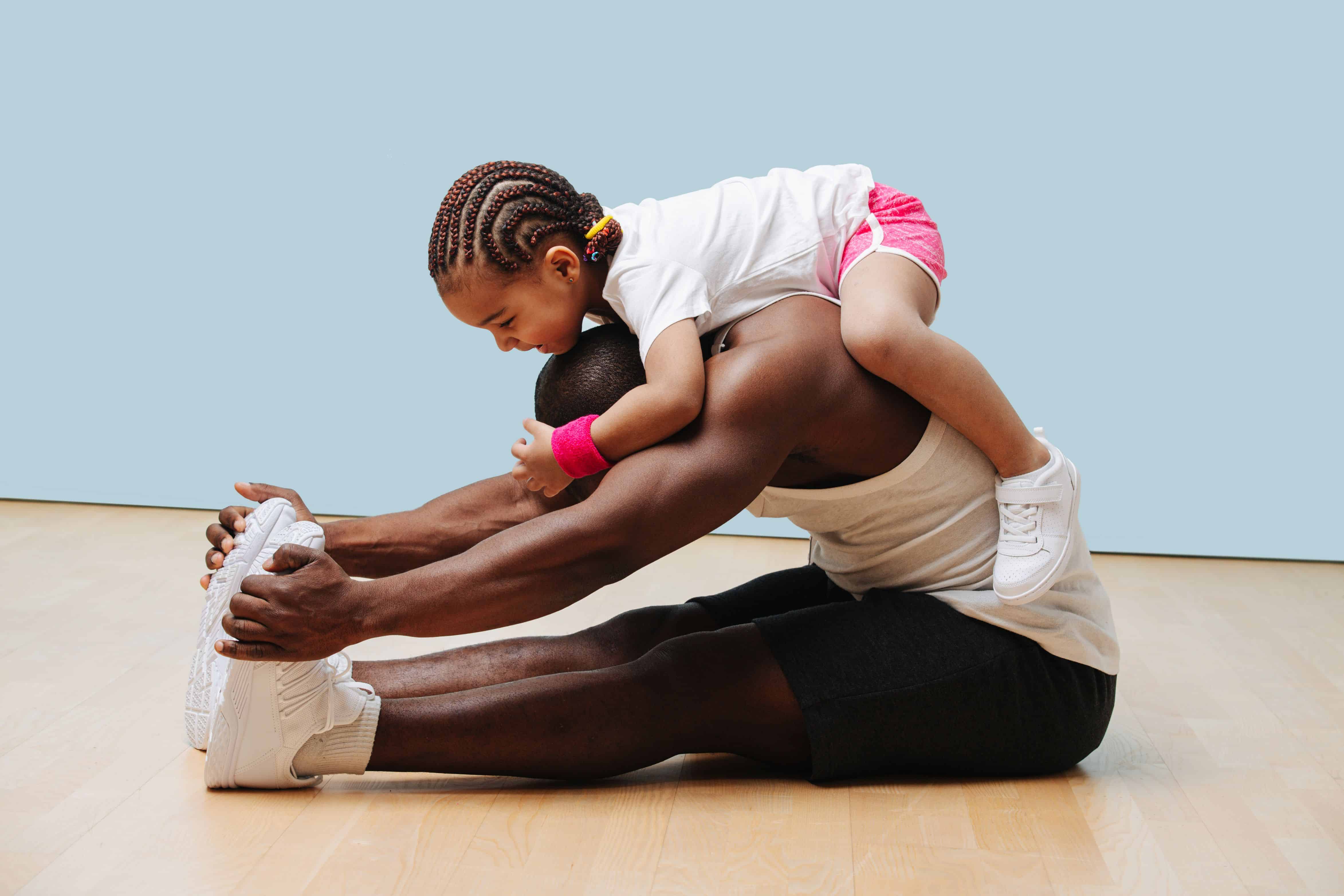 Daughter climbes on her father's neck as he stretches his back and knees on the floor. They having fun, entertaining themselves in isolation.