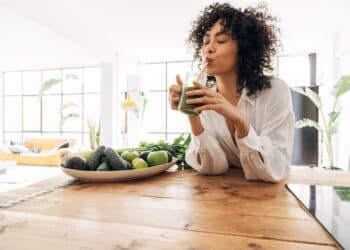 Young african american woman drinking green juice with reusable bamboo straw in loft apartment. Home concept. Healthy lifestyle concept. Copy space