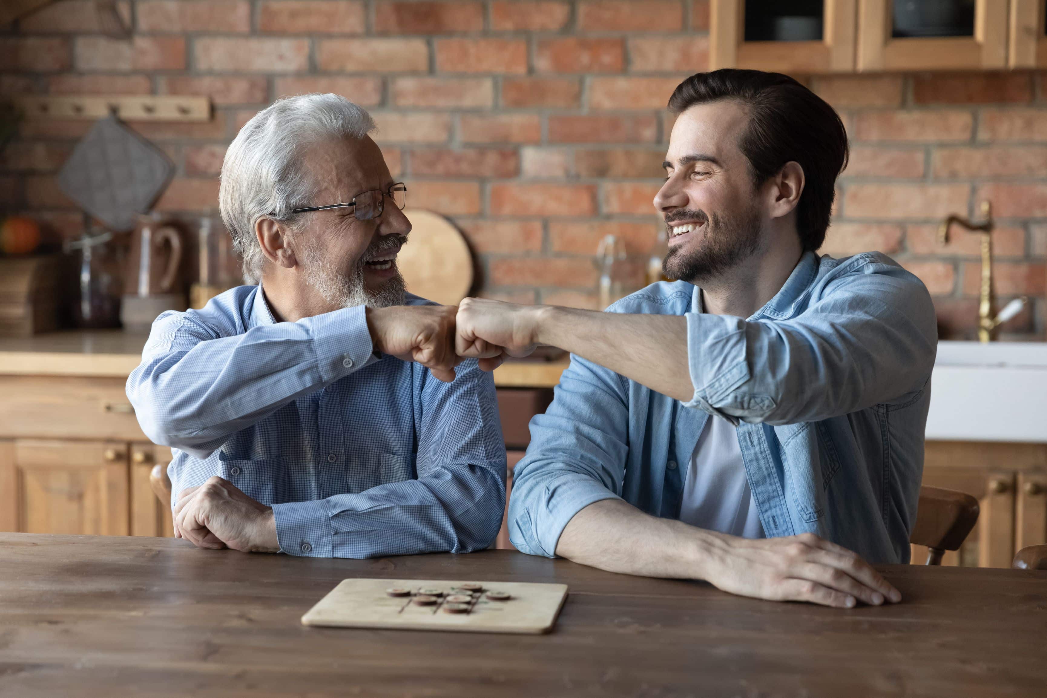Happy elderly dad and grownup son giving fist bumps, making friends gesture, celebrating achievement in boardgame, expressing respect, playing checker draughts, enjoying hobby, leisure time