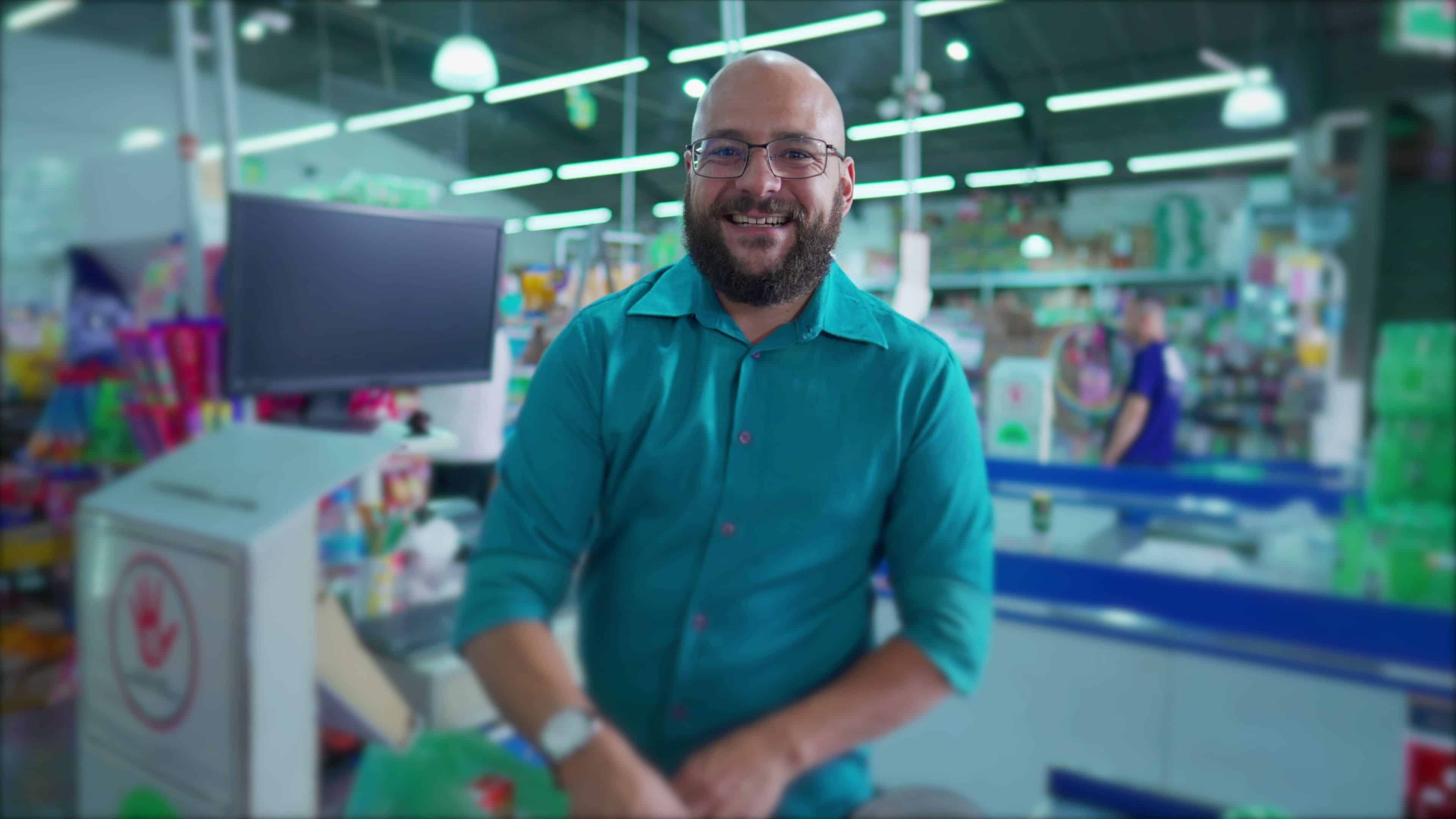 One happy Brazilian employee of supermarket at checkout scanning products and putting into plastic bags while smiling at camera. Grocery Market scene in South America