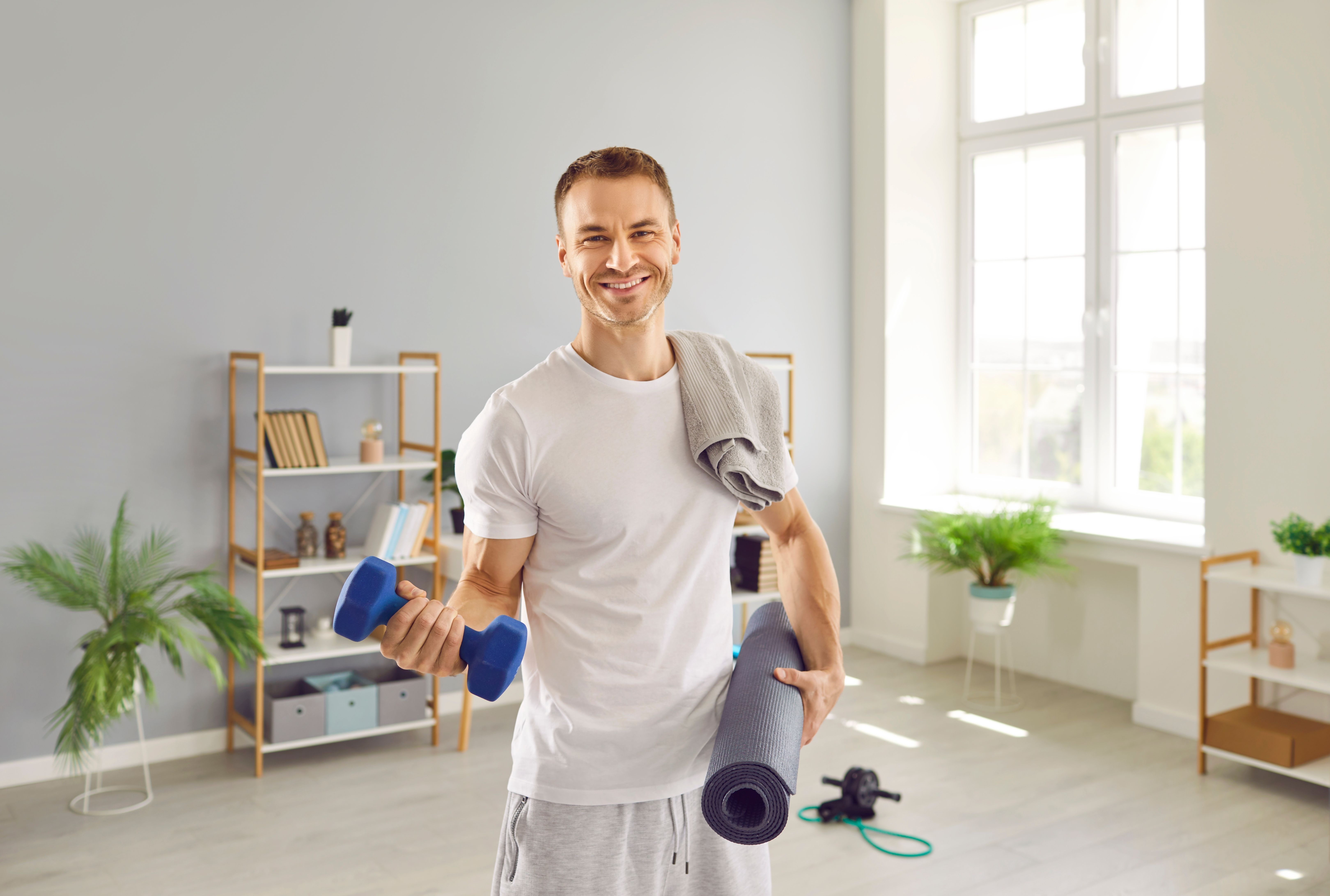 Portrait happy strong fit healthy young man in white T shirt, with towel on shoulder, standing in living room at home, holding fitness dumbbells and sports workout mat, looking at camera, and smiling