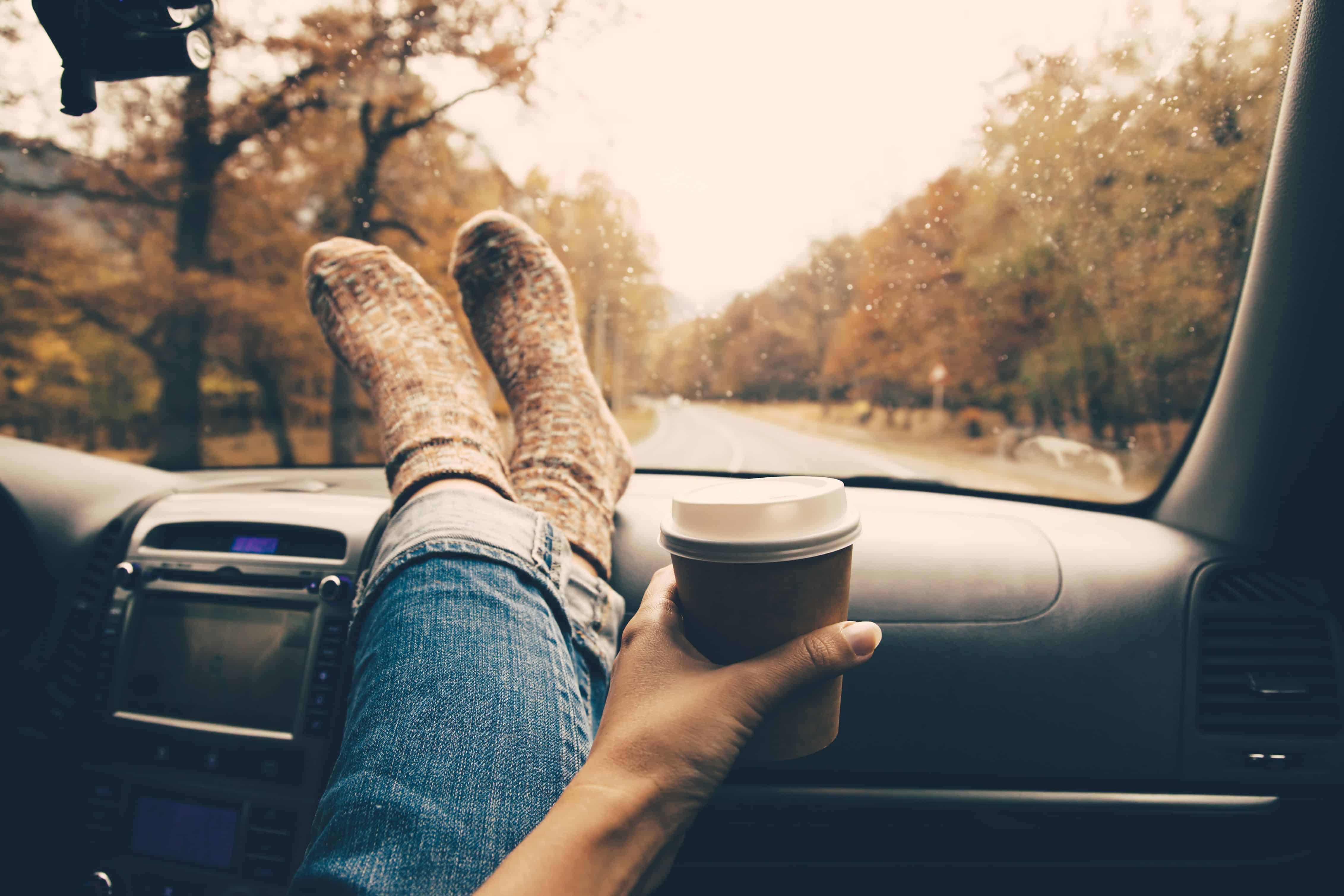 Woman feet in warm socks on car dashboard. Drinking take away coffee on road. Fall trip. Rain drops on windshield. Freedom travel concept. Autumn weekend.
