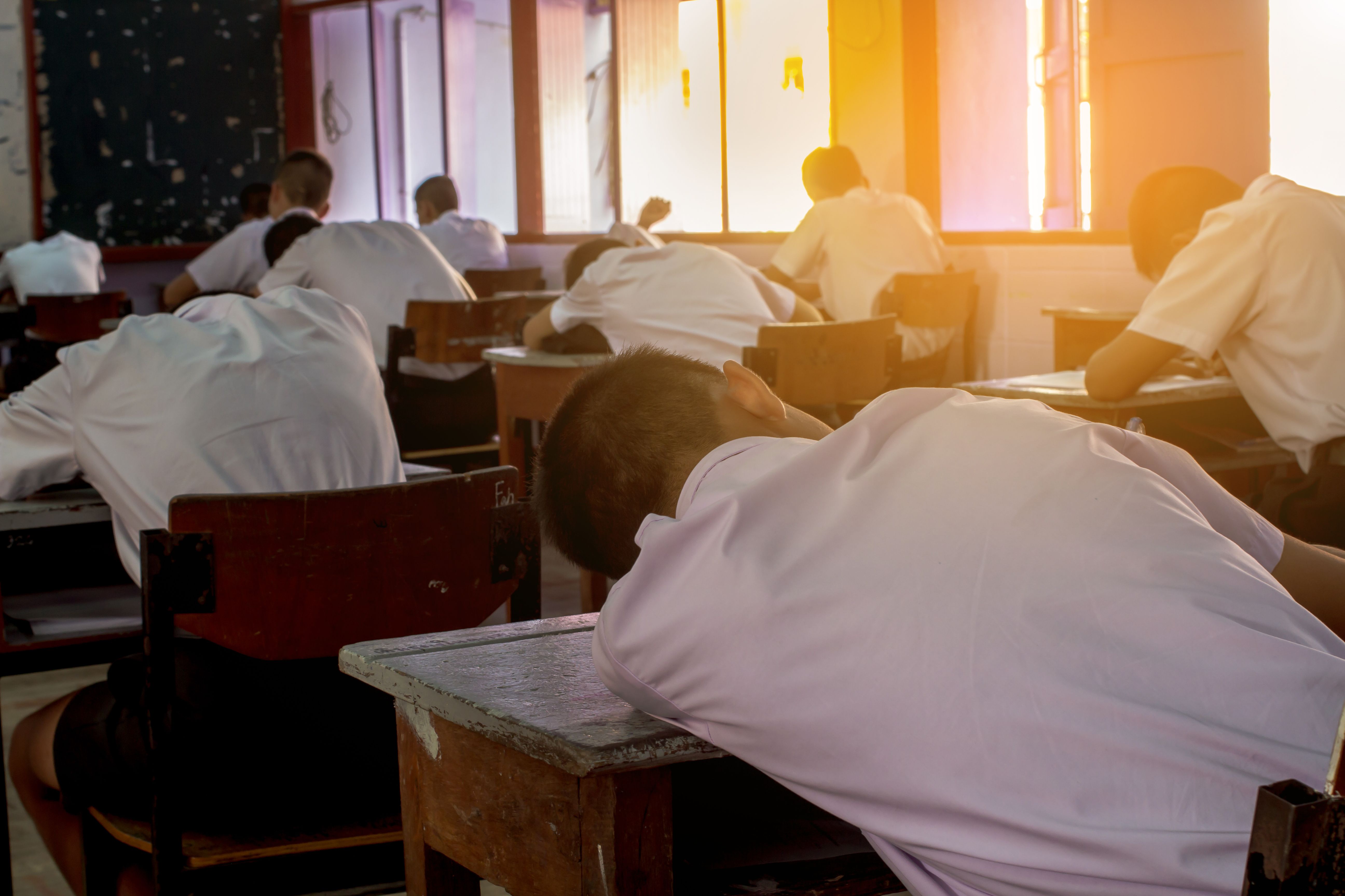 Poor Student with student’s uniform so tired of the exam. Lying down on the arm in the classroom. Sleepy student fall asleep during test at noon in the hot season.