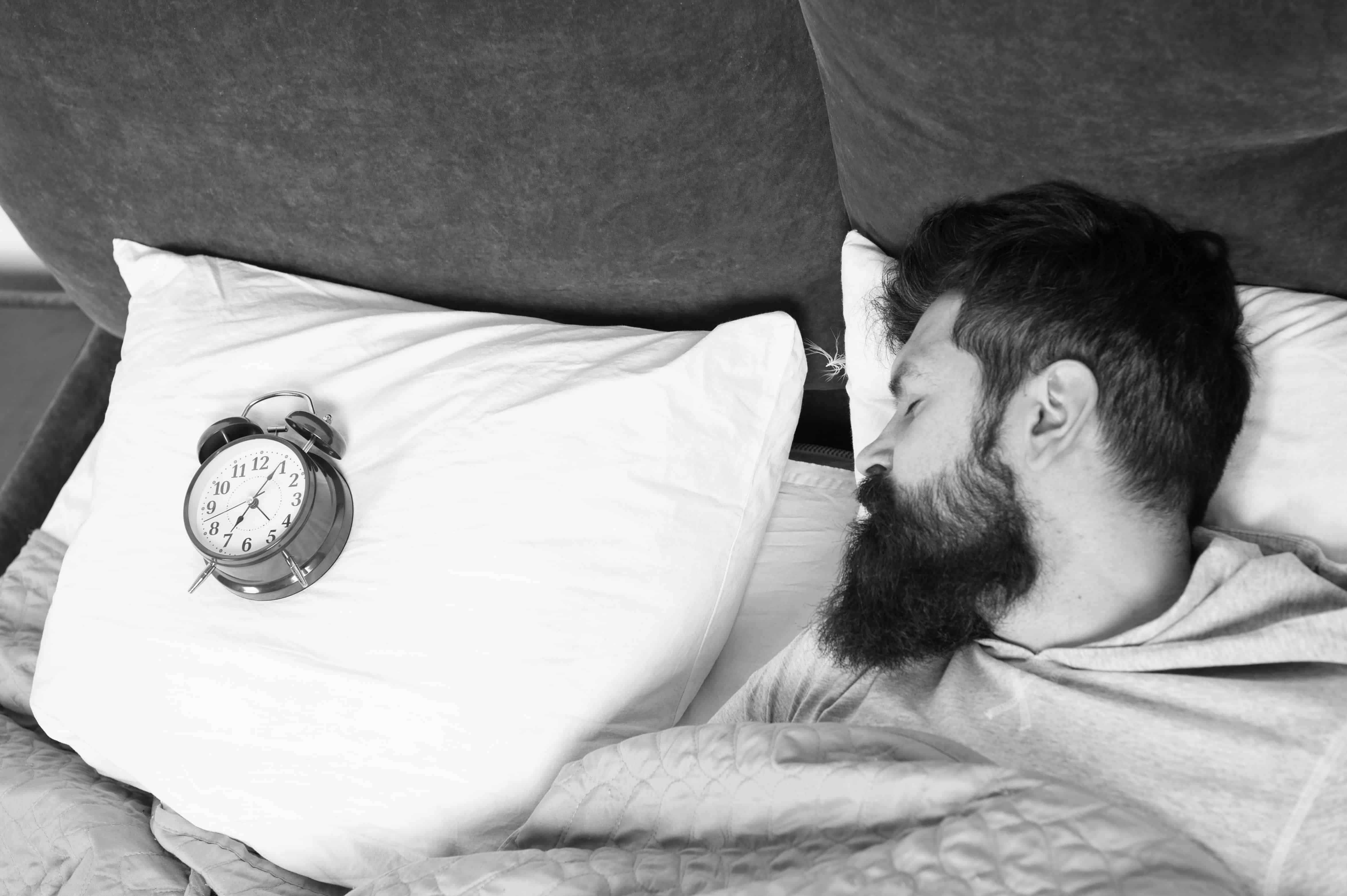 Man bearded hipster sleepy face in bed with alarm clock. Problem with early morning awakening. Get up with alarm clock. Overslept again. Tips for waking up early. Tips for becoming an early riser.