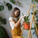 Young smiling woman gardener in glasses wearing overalls, taking care for orchid in old red milk can standing on orange vintage ladder. Home gardening, love of houseplants, freelance.
