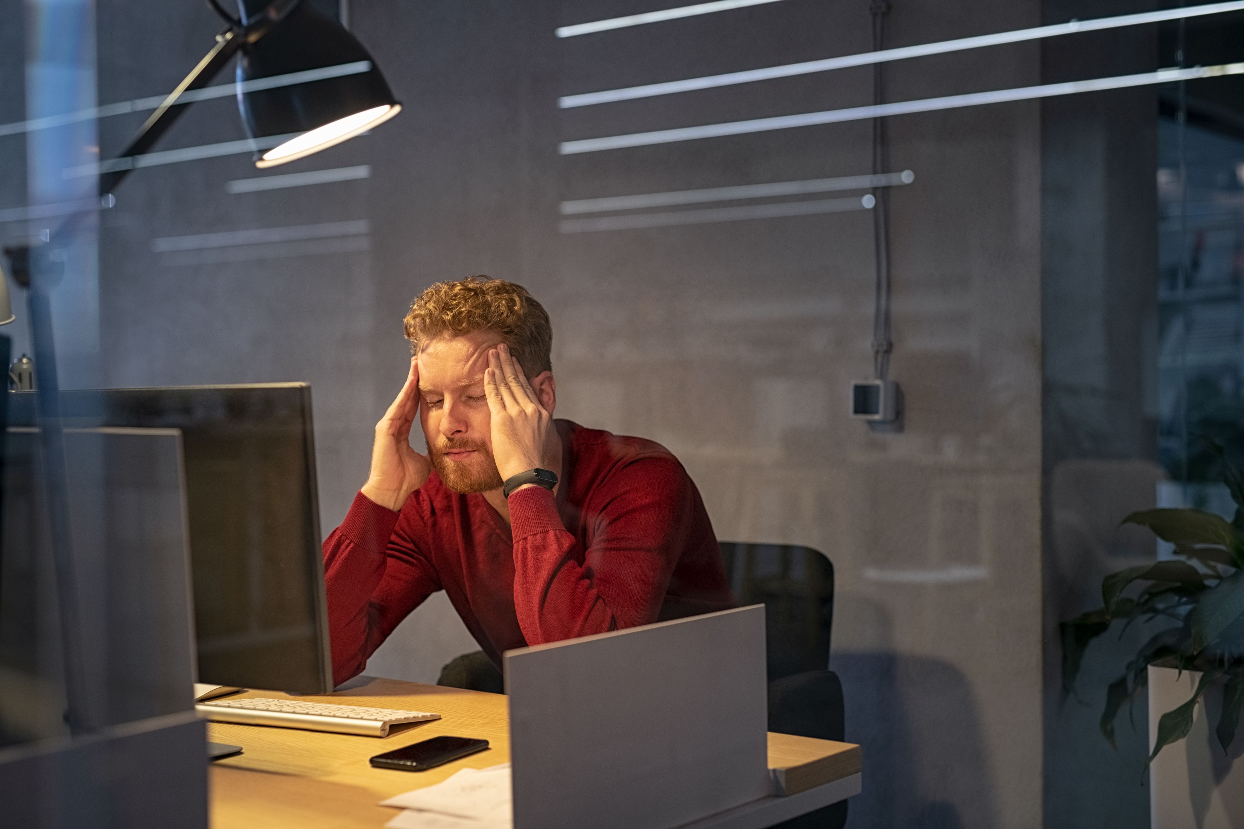 Upset businessman at desk with terrible headache. Tired business man working late in office. Young stressed man feeling headache after hard working for long hours on computer, overwork and burnout.