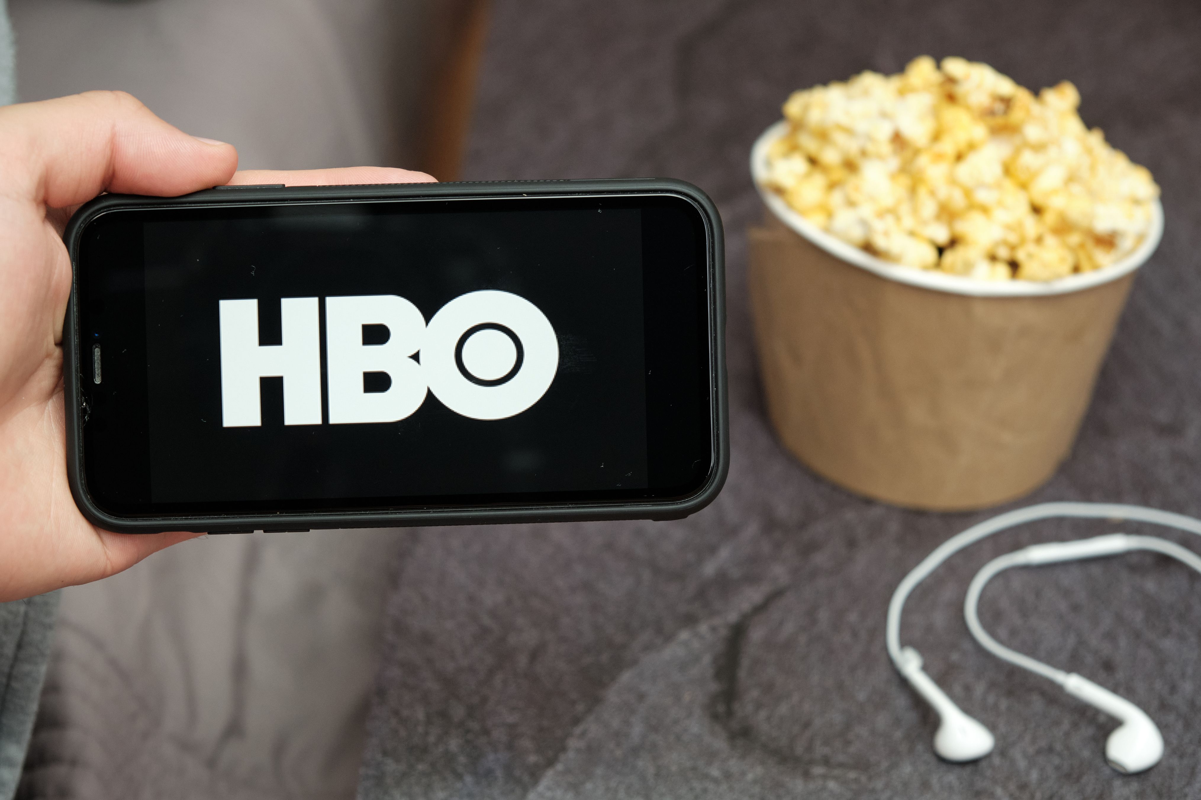 Close up mans hand holding a mobile phone with HBO logo with Apple earphones and popcorn box next to him, August 2020, San Francisco, USA