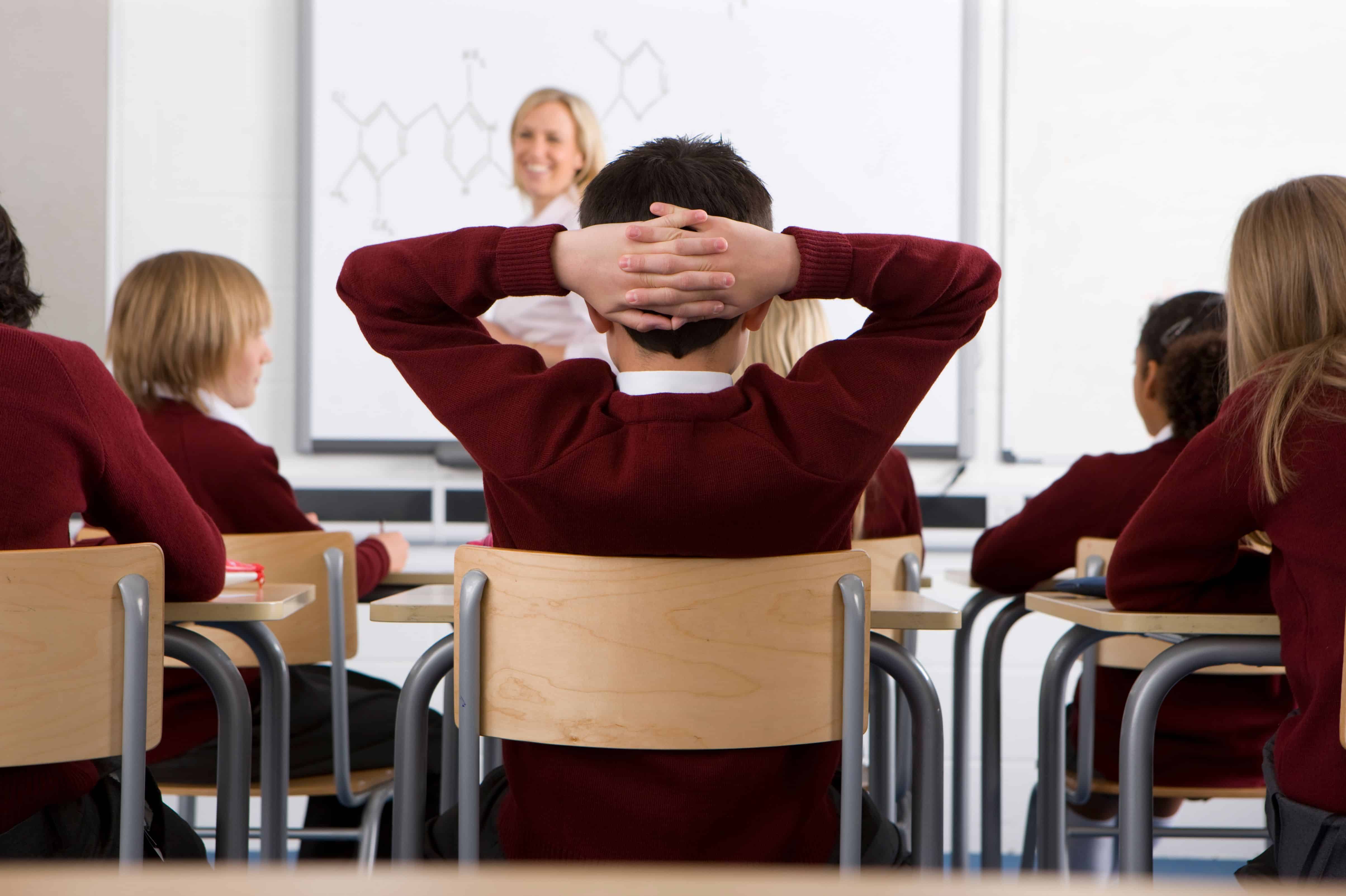 Rear view of a school boy with his hands behind head while listening to the subject being taught by the teacher in a classroom